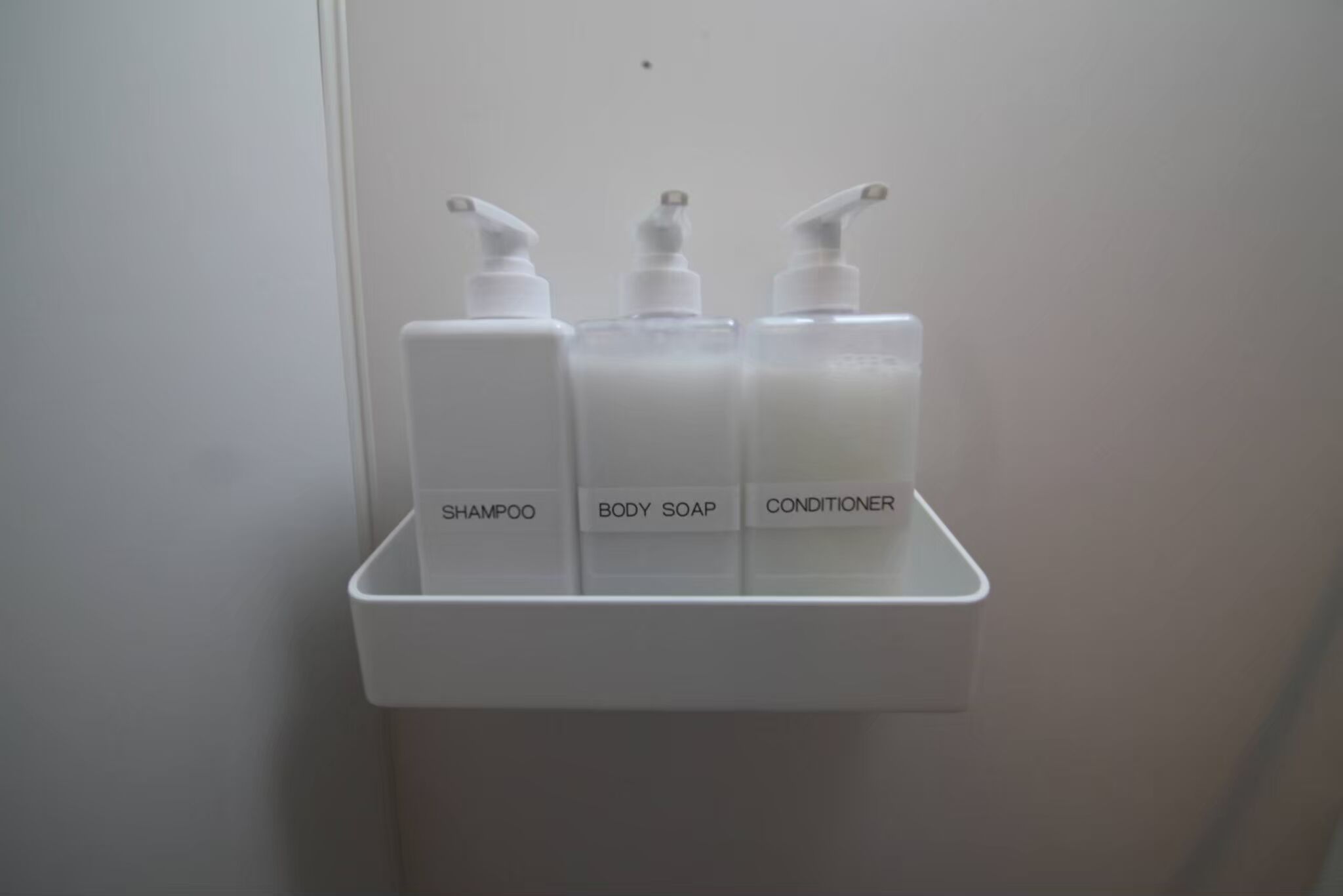 Een witte plank met daarboven drie flessen, gelabeld als shampoo, douchezeep en conditioner.