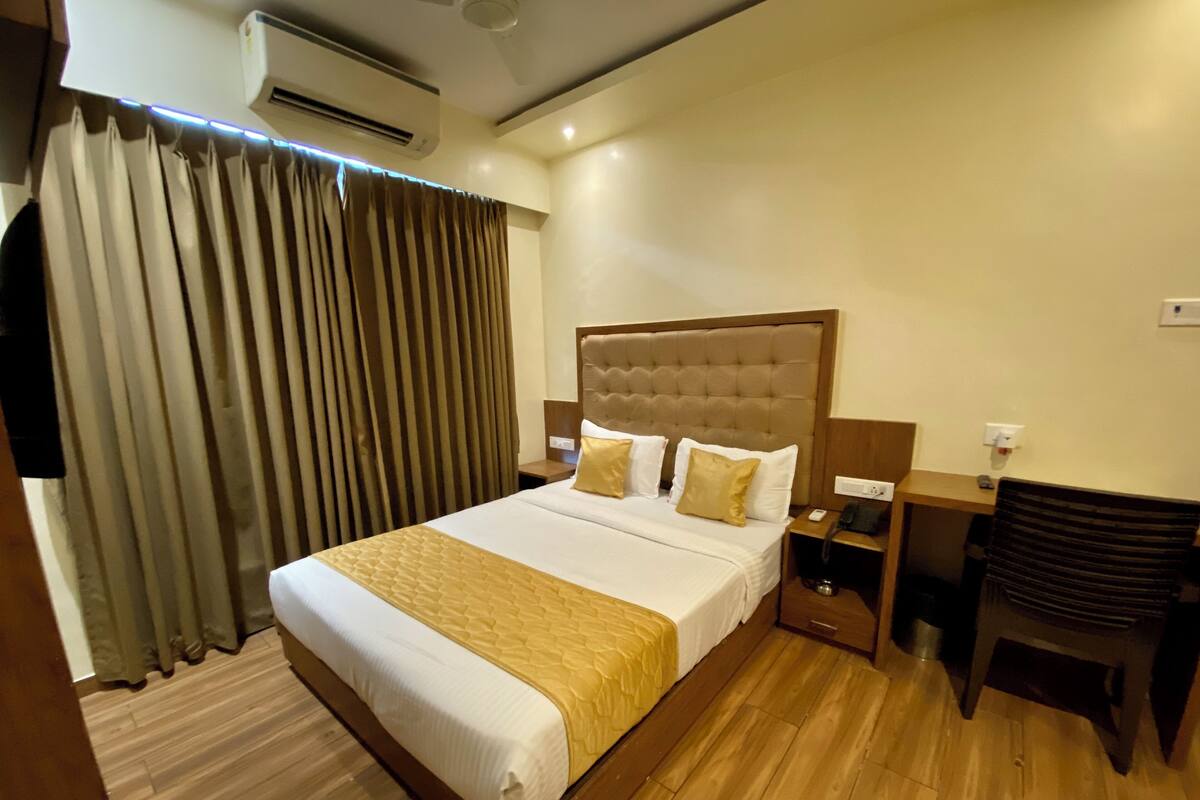 A hotel room with a large bed, a wooden headboard, a bedside table with a lamp, a chair, and a window with curtains.