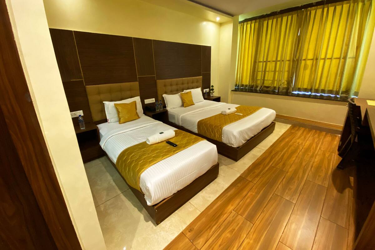 A hotel room with two beds, wooden flooring, a large window with yellow curtains, and a ceiling fan.