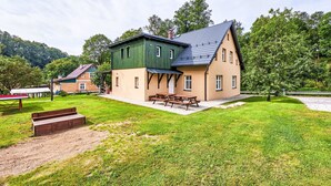 Une maison à deux étages, avec une façade verte et un rez-de-chaussée de couleur beige, dotée d’un patio comprenant une table de pique-nique et des bancs.