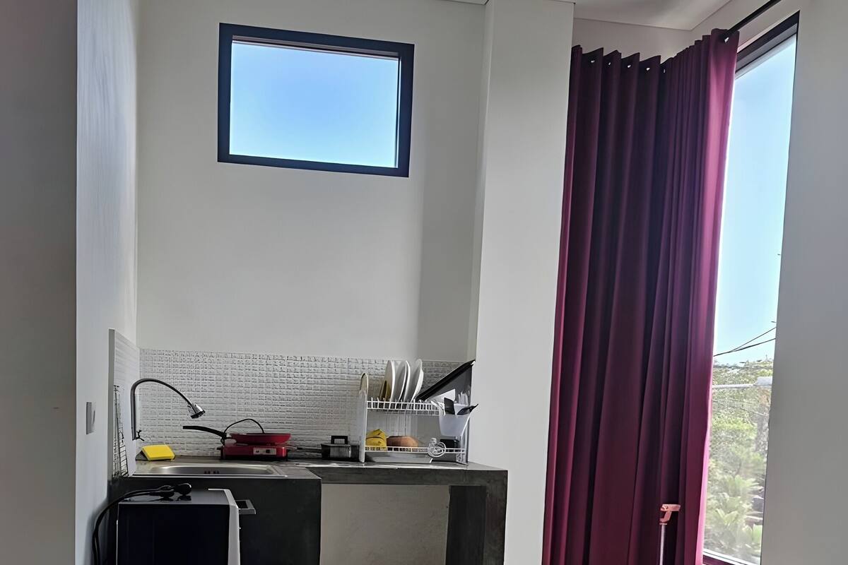 A hotel room with a bed, a desk with a computer, a small kitchen area, and a window with curtains.