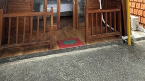 Una pequeña cabaña de madera tipo A con un porche cubierto y una cama individual visible en el interior.