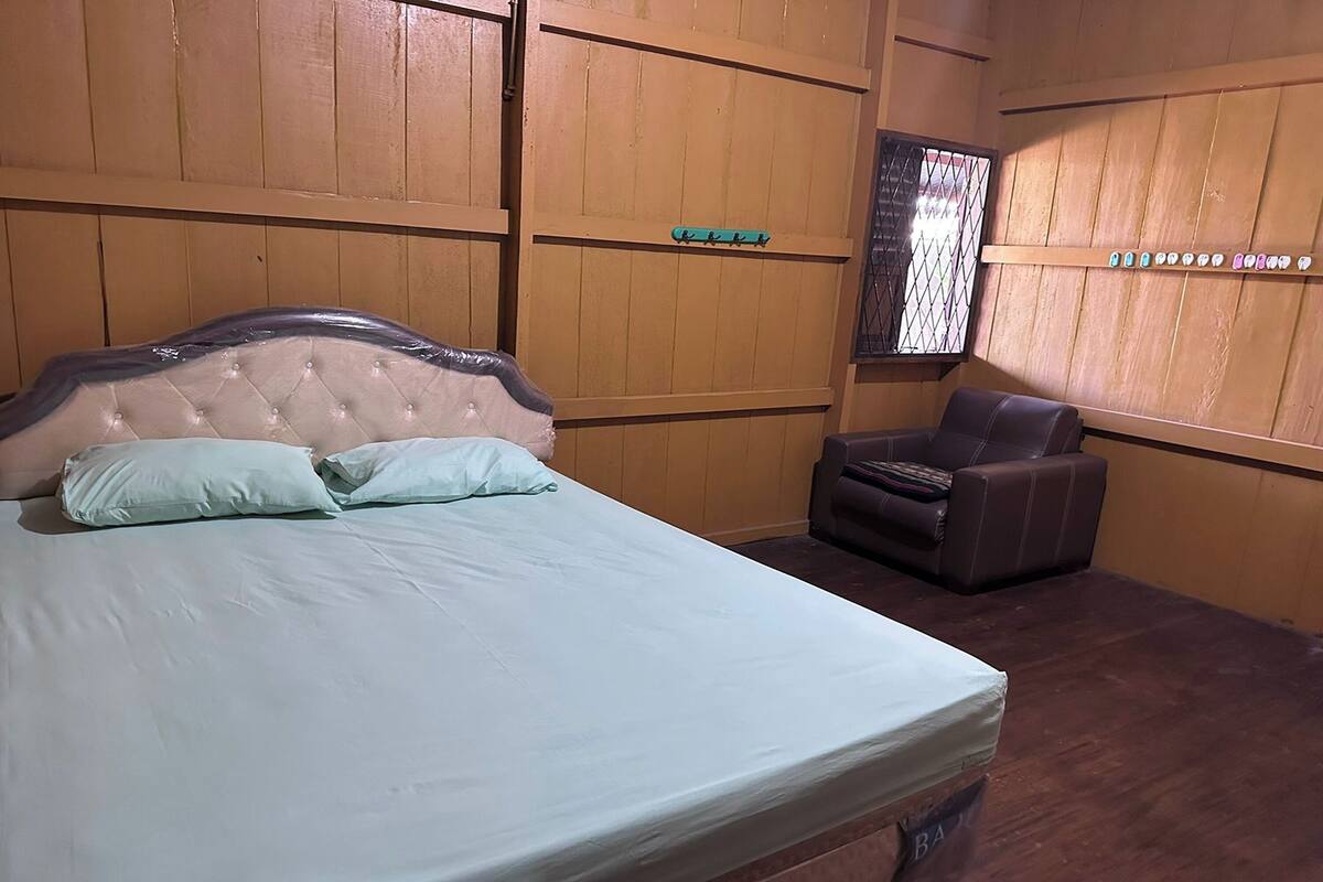 A bedroom with a bed, a chair, wooden panel walls, and a window with a metal grid.