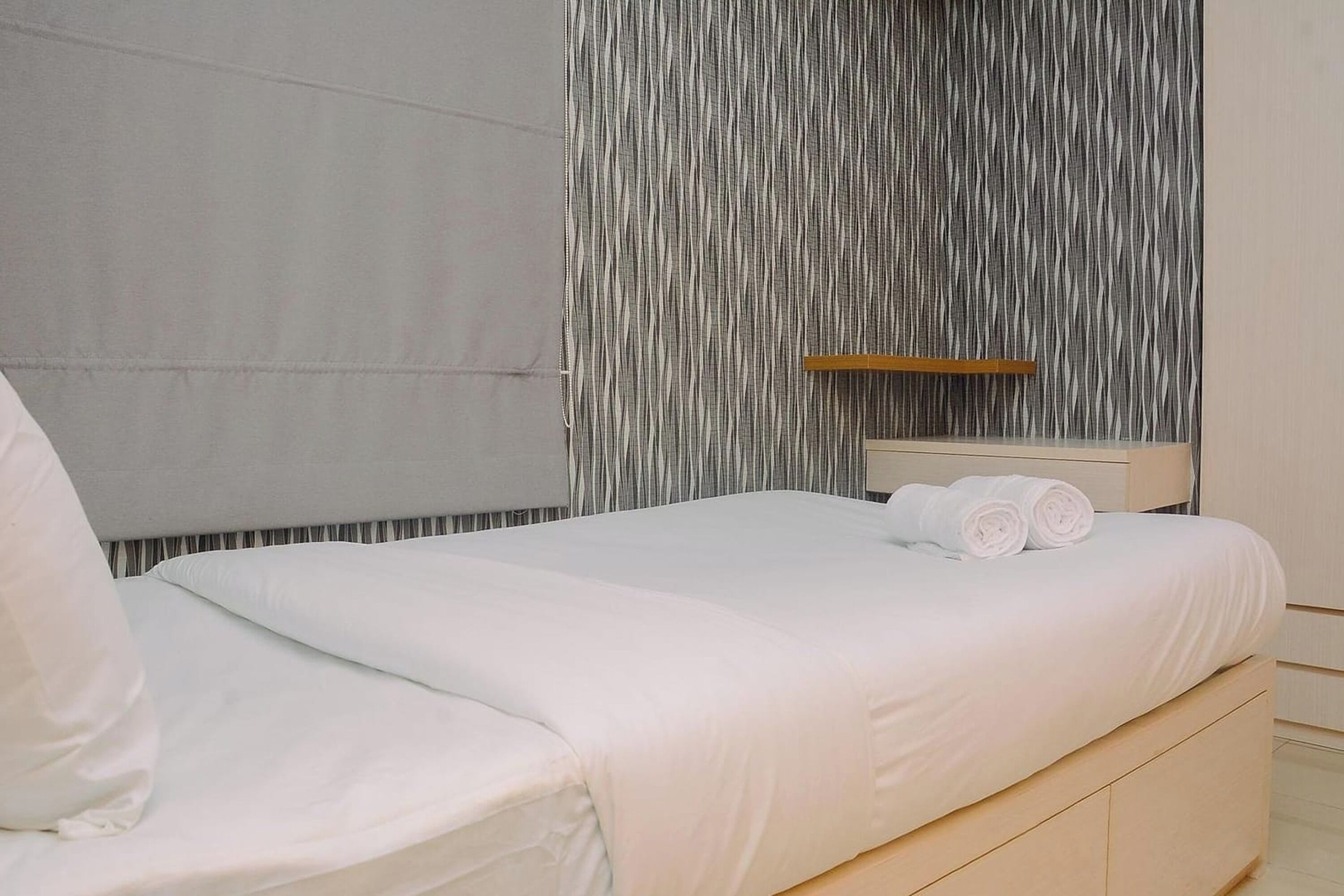 A neatly made bed with white linens and rolled towels, a wooden headboard, and a textured wall behind.