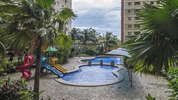 A swimming pool area with a slide, surrounded by palm trees and a playground.