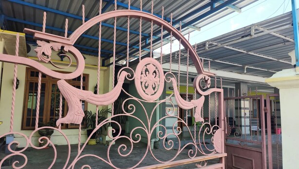 A pink metal gate with intricate designs, a covered area with a corrugated metal roof, and a building with a green facade and windows.