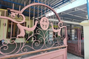 Ein pinkfarbenes Metallgitter mit aufwendigen Verzierungen, ein überdachter Bereich mit Wellblechdach und ein Gebäude mit grüner Fassade und Fenstern.