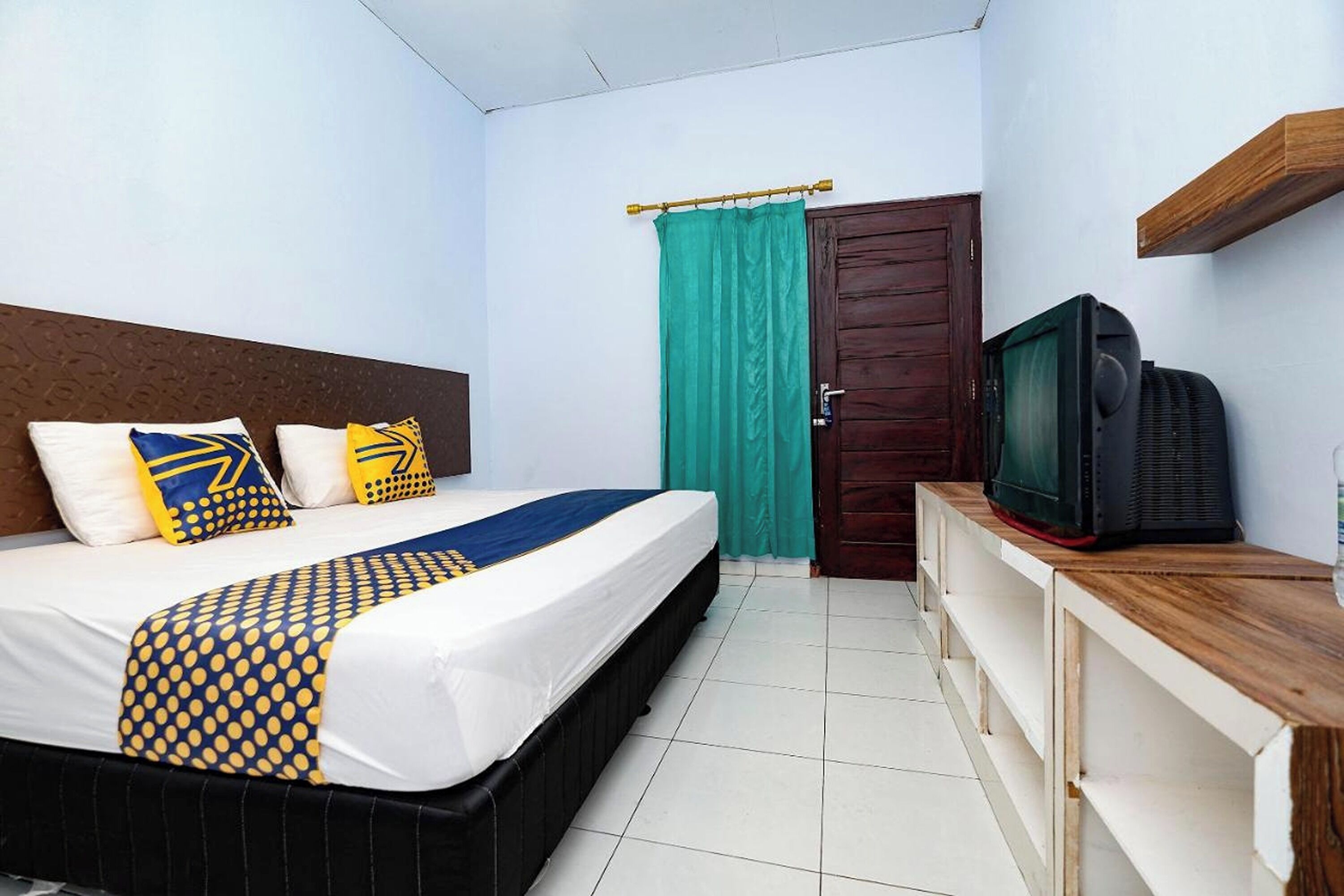 A bedroom with a bed, a television on a wooden cabinet, a door with a curtain, and a shelf above the bed.