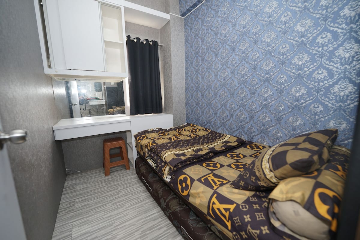 A compact bedroom with a bed, a small stool, a white desk with a mirror, and patterned wallpaper.