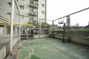 Una cancha de baloncesto al aire libre rodeada por una cerca de malla metálica, con un edificio al fondo.
