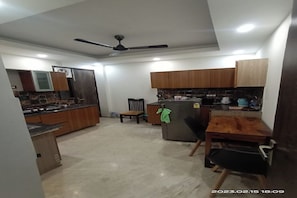 A modern kitchen with wooden cabinets, a refrigerator, a dining table with chairs, and a ceiling fan. - Mintstar Apartment And Suites, East Of Kailash (New Delhi)