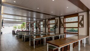 Een ruime eetruimte met lange houten tafels en witte stoelen, grote ramen met decoratief glas en een plafond met inbouwverlichting.