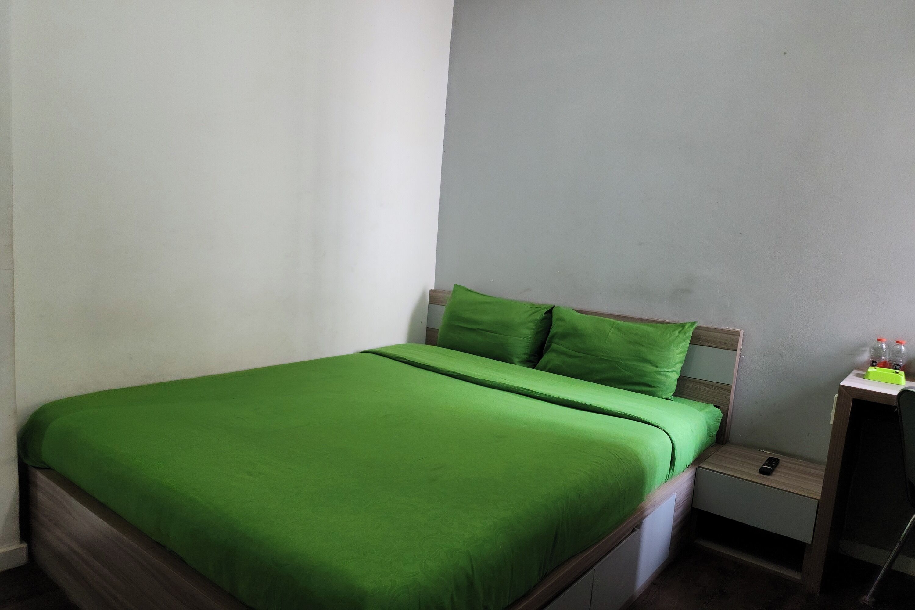 A single-bed room with a green bedspread, a wooden headboard, a nightstand with a phone, and a small table with bottles.