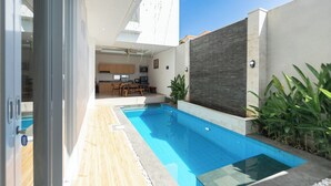 Una piscina esterna moderna con un solarium in legno e una zona cucina visibile sullo sfondo.