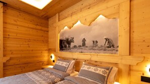 Habitación de cabaña de madera con dos camas, un cuadro grande en la pared con una escena campestre y luces empotradas.