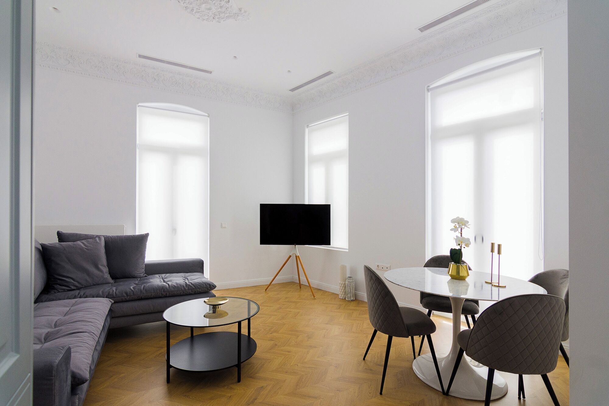 Un salon moderne avec un canapé gris, une table basse en verre, une table à manger ronde avec des chaises, et un téléviseur à écran plat fixé au mur.