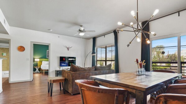 A spacious living area with a large wooden dining table, leather chairs, a ceiling fan, and a TV displaying streaming services.