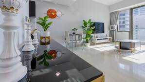 A modern living room with a large window, a flat-screen TV, and a dining area with a white table and chairs. - Gorgeous 1 Bed l City Views l near Media City Park (Dubai Marina)