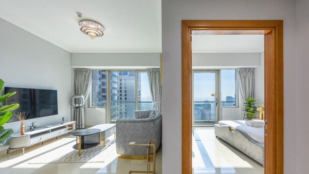 A modern living room with a large TV, a grey sofa, a small round table, and a balcony with a city view. - Gorgeous 1 Bed l City Views l near Media City Park (Dubai Marina)