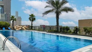 A swimming pool with clear blue water, surrounded by palm trees and modern buildings in the background. - Gorgeous 1 Bed l City Views l near Media City Park (Dubai Marina)