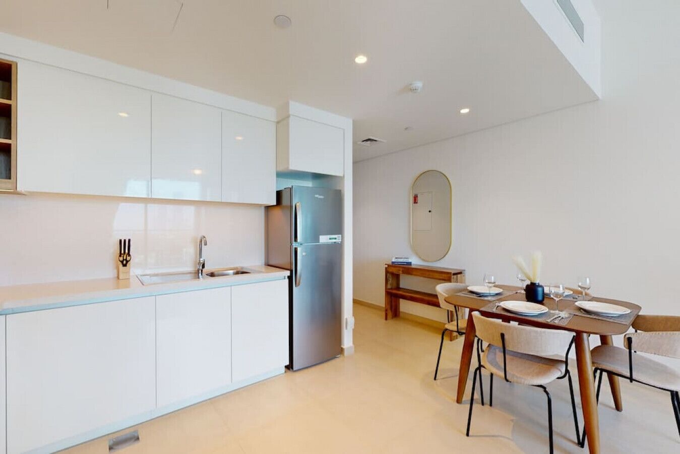 Cocina moderna con armarios blancos, un refrigerador de acero inoxidable y una zona de comedor con una mesa y sillas de madera.