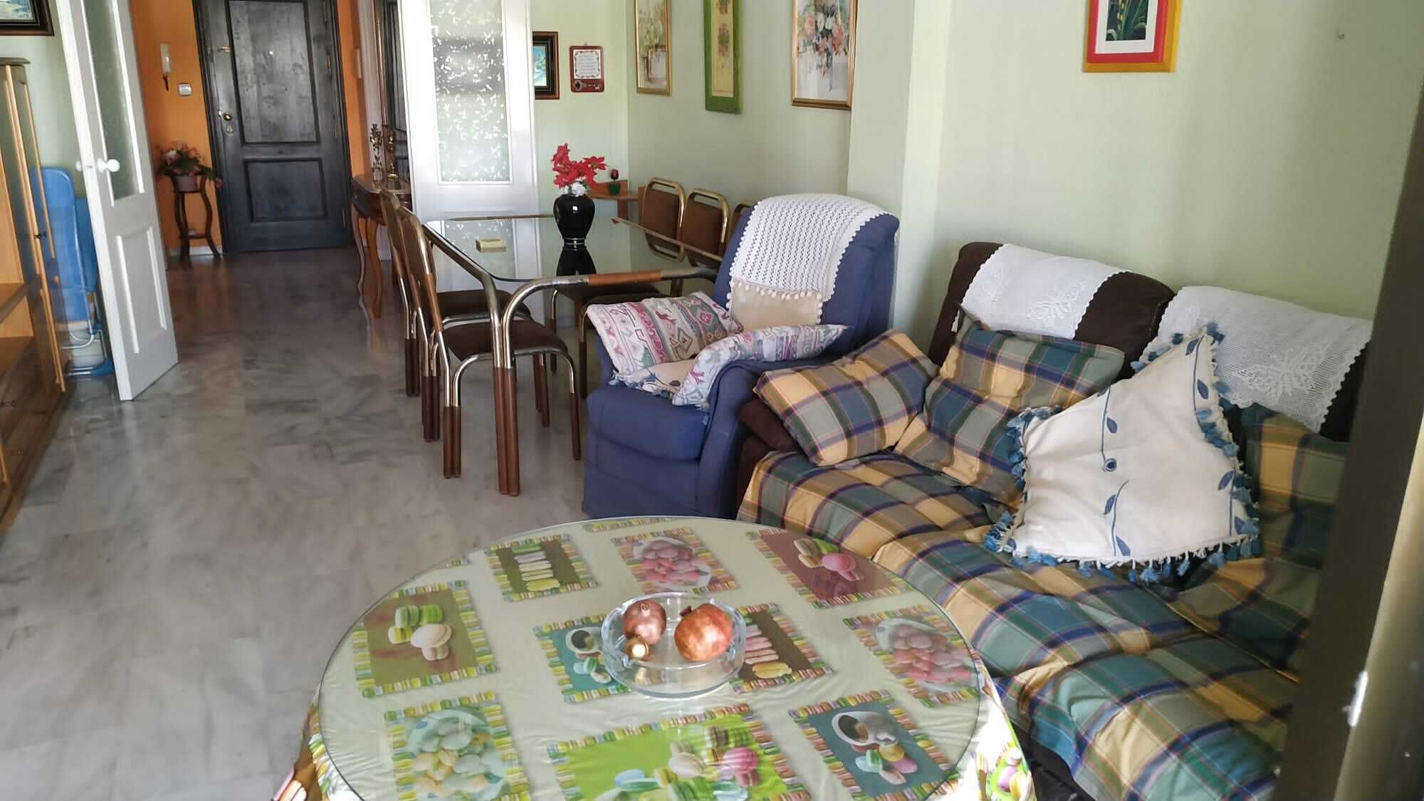 Un salon avec un canapé bleu, une table basse ronde recouverte d’une nappe à motifs floraux, et un coin repas avec une table et des chaises en bois.