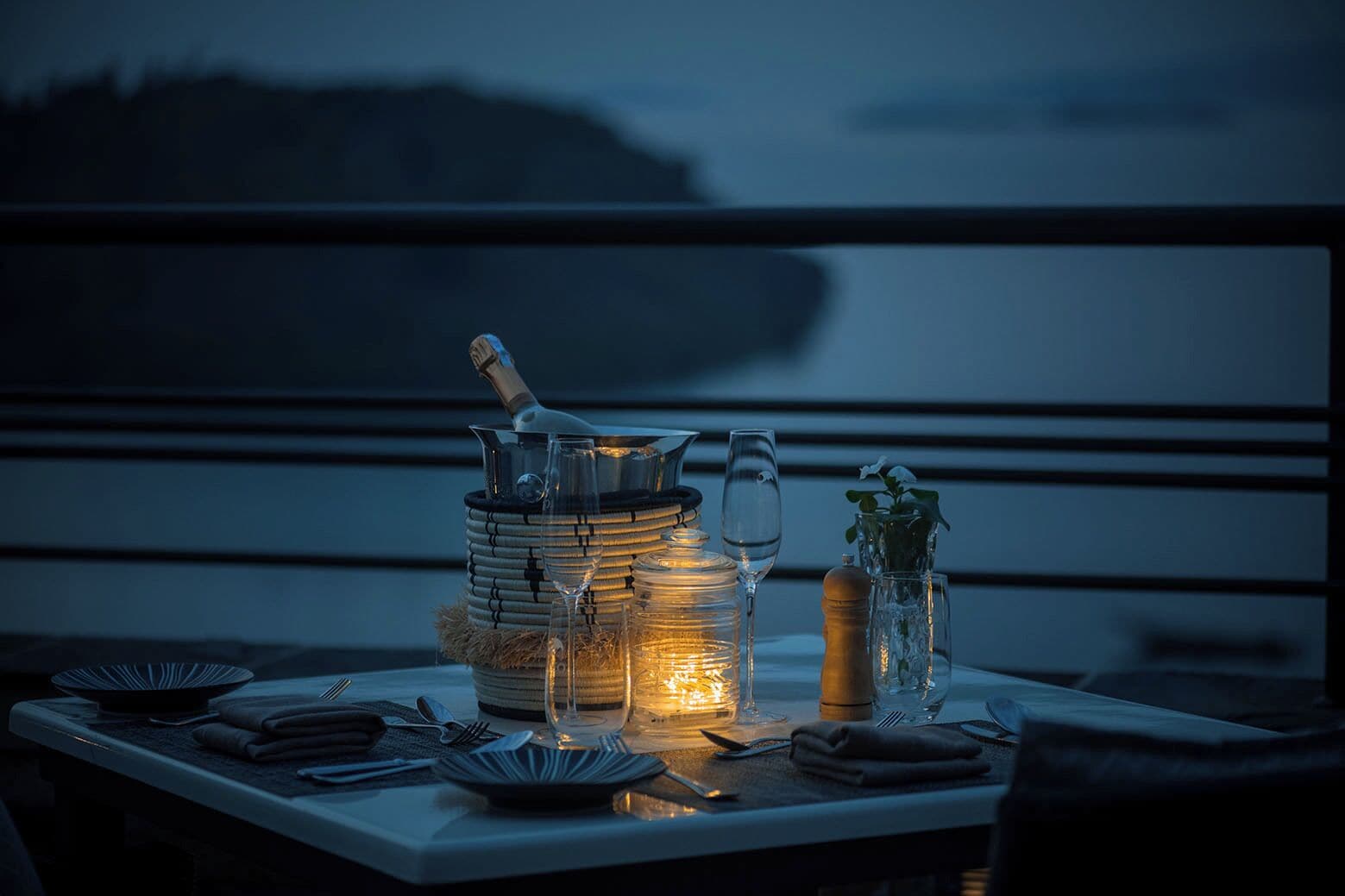 A table set for dining with a candle, a glass pitcher, and a bottle, overlooking a body of water.