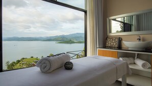 Habitación de hotel con un ventanal grande que da a un lago y montañas, una cama con una toalla enrollada, un pequeño cuenco decorativo y un lavabo blanco con un espejo encima.