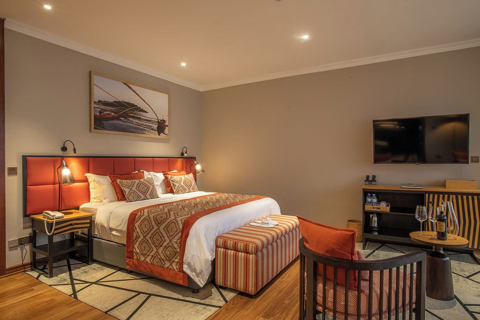 Una habitación de hotel con cama, mesitas de noche, televisión, zona de estar con un sillón y reposapiés, y un cuadro en la pared.
