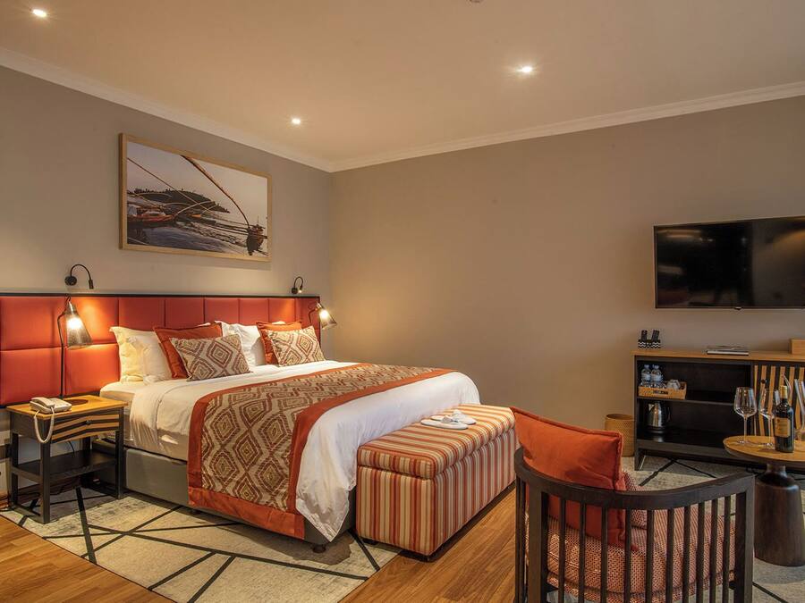 Una habitación de hotel con cama, mesitas de noche, televisión, zona de estar con un sillón y reposapiés, y un cuadro en la pared.