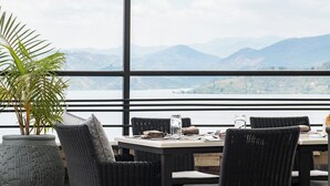 A dining area with wicker furniture, a potted plant, and a table set for two with a view of mountains and a lake. - LUX* Lake Kivu (Bwishyura)