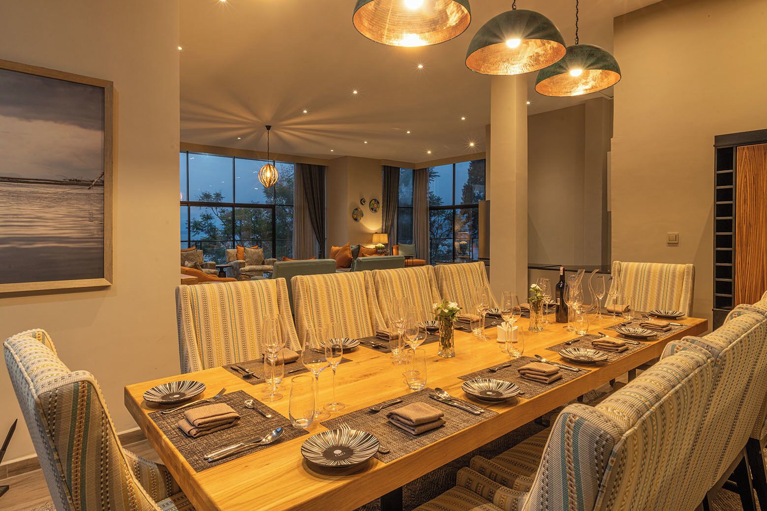 Uma sala de jantar com uma mesa de madeira comprida posta para uma refeição, cadeiras estofadas e iluminação pendente.