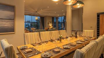 Uma sala de jantar com uma mesa de madeira comprida posta para uma refeição, cadeiras estofadas e iluminação pendente.
