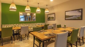 Un comedor con una mesa grande, varias sillas, lámparas colgantes, una pared verde y un cuadro enmarcado.