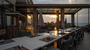 Un espacio de comedor al aire libre con una mesa larga, sillas y sombrillas, con vista a un cuerpo de agua.