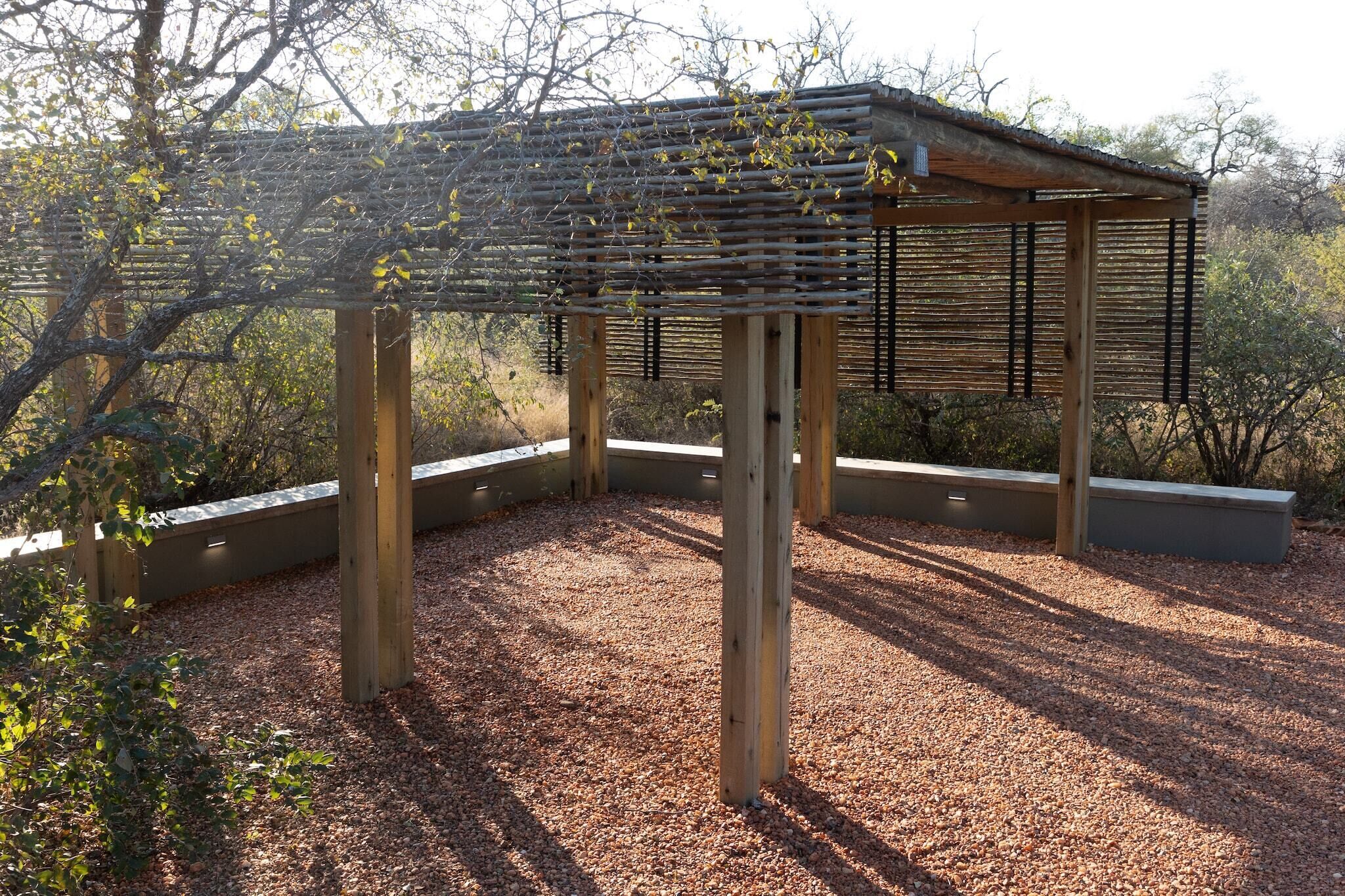 Une structure de pergola en bois, surmontée d’un toit en métal, entourée de gravier et de plantes, dans un cadre extérieur.