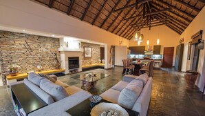 A spacious living area with a stone wall, a fireplace, and a modern kitchen in the background. - Luxury villa with a swimming pool in Hoedspruit (Hoedspruit)