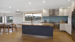A modern kitchen with a central island, a dining area with wooden chairs, and a large window offering a view of the outdoors. - Bella Vista Valley Views (BLACKHEATH)