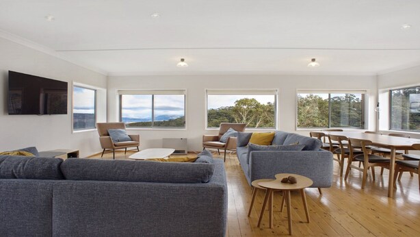 A modern living room with a large grey sofa, a smaller grey sofa, and a dining table with chairs. There are large windows with a view of the outdoors. - Bella Vista Valley Views (BLACKHEATH)