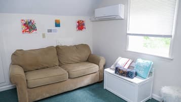 Een kamer met een beige bank, een witte bijzettafel met boeken en een kussen, een raam met jaloezieën en drie abstracte schilderijen aan de muur.