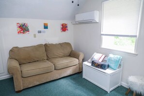 Ein Zimmer mit einem beigen Sofa, einem weißen Nachttisch mit Büchern und einem Kissen, einem Fenster mit Jalousien und drei abstrakten Bildern an der Wand.