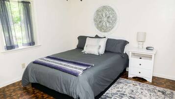 A bedroom with a bed, a nightstand, a lamp, a window with curtains, and a decorative wall piece.