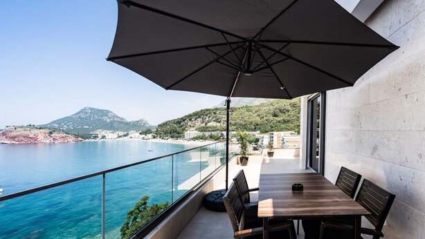 A balcony with a table and chairs, overlooking a coastal view with a mountain in the distance. - Hotel G Star (Sutomore)