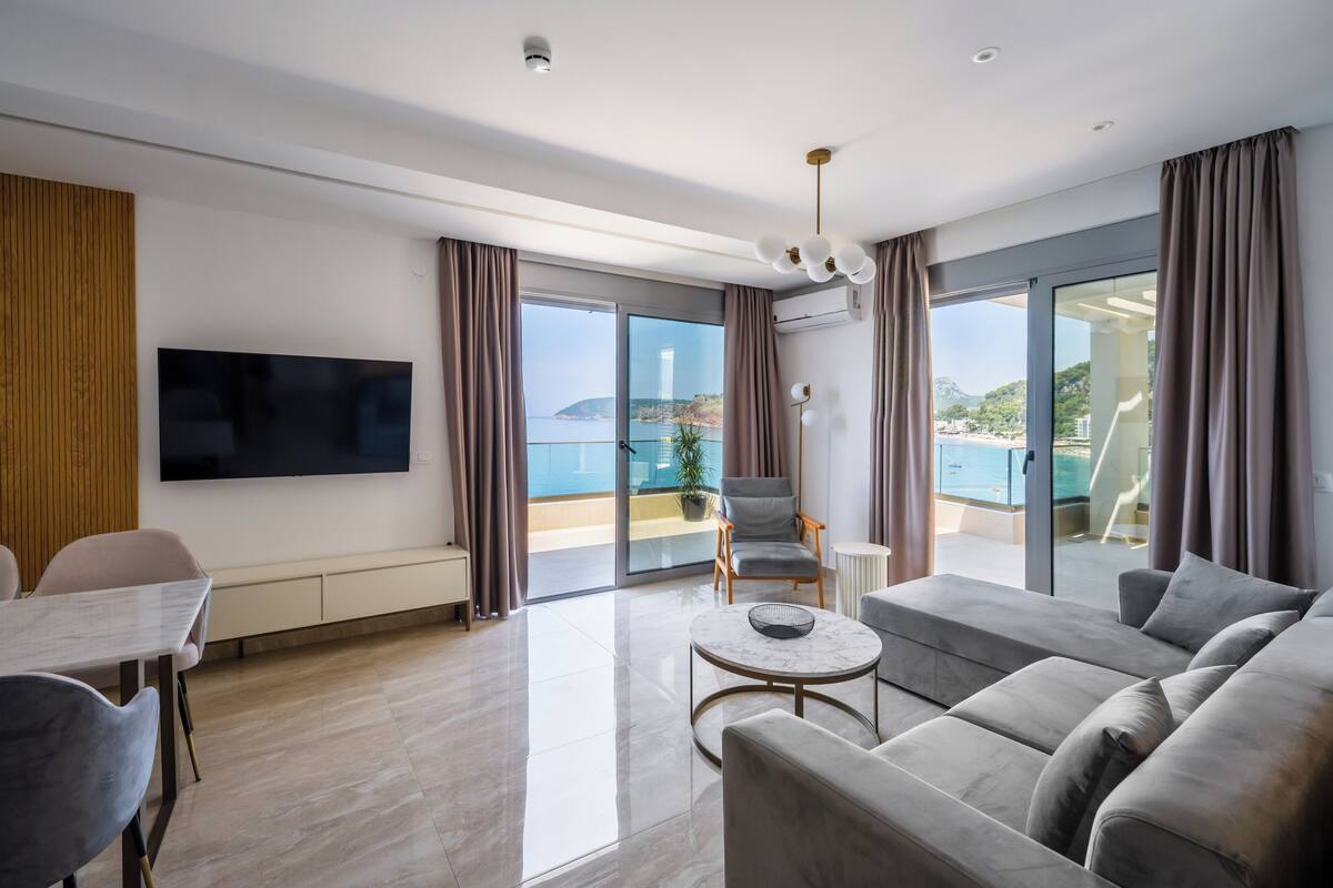 A modern living room with a flat-screen TV, a sofa, a dining table, and a balcony with a view of the sea.