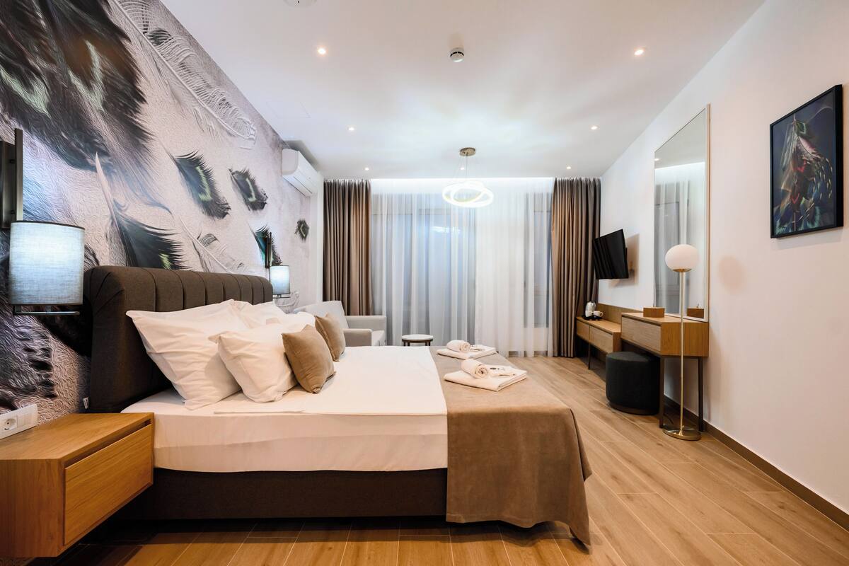 A modern hotel room with a large bed, a desk, a chair, a TV, and a large window with curtains.