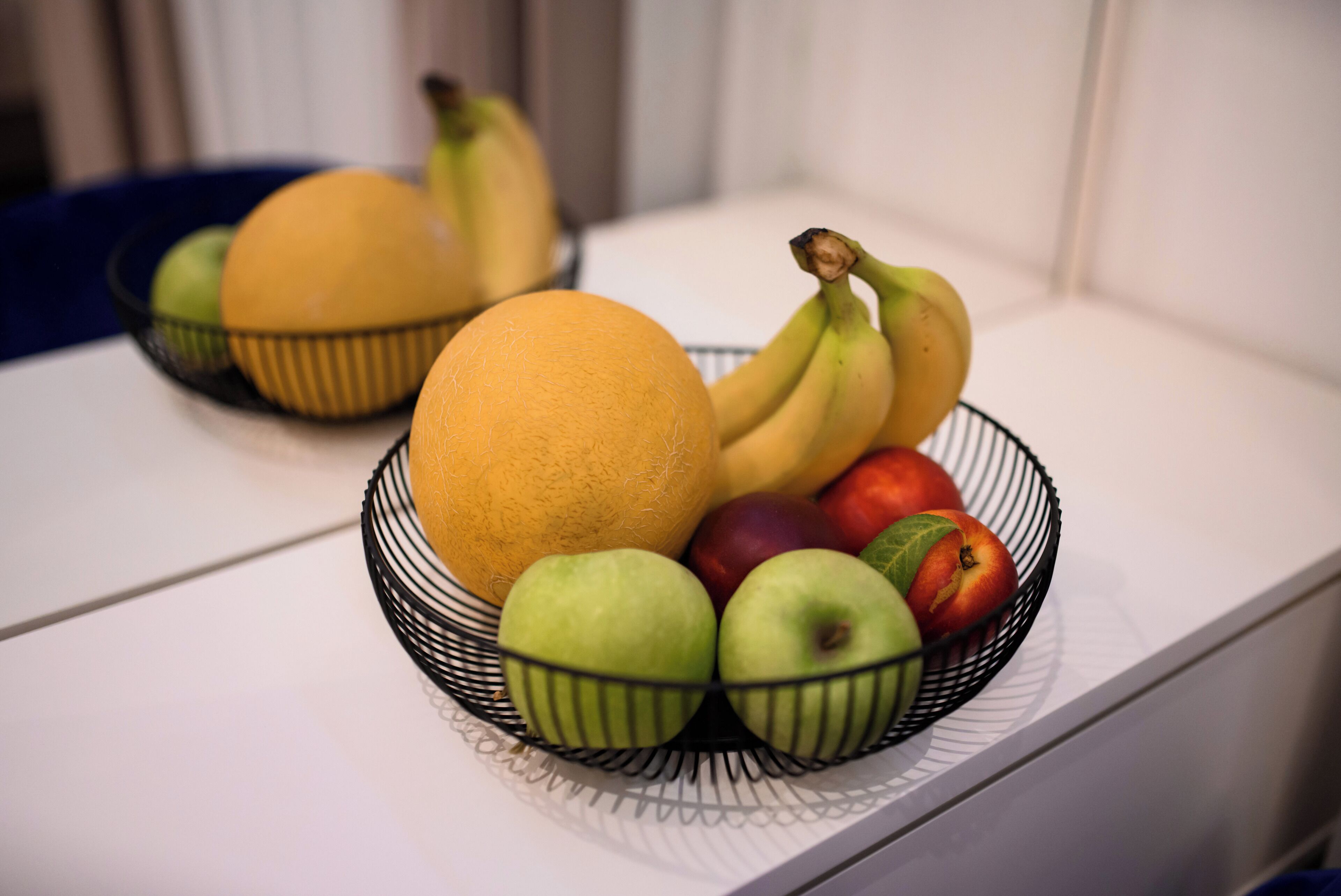 A fruit bowl containing apples, bananas, and a melon on a white surface.