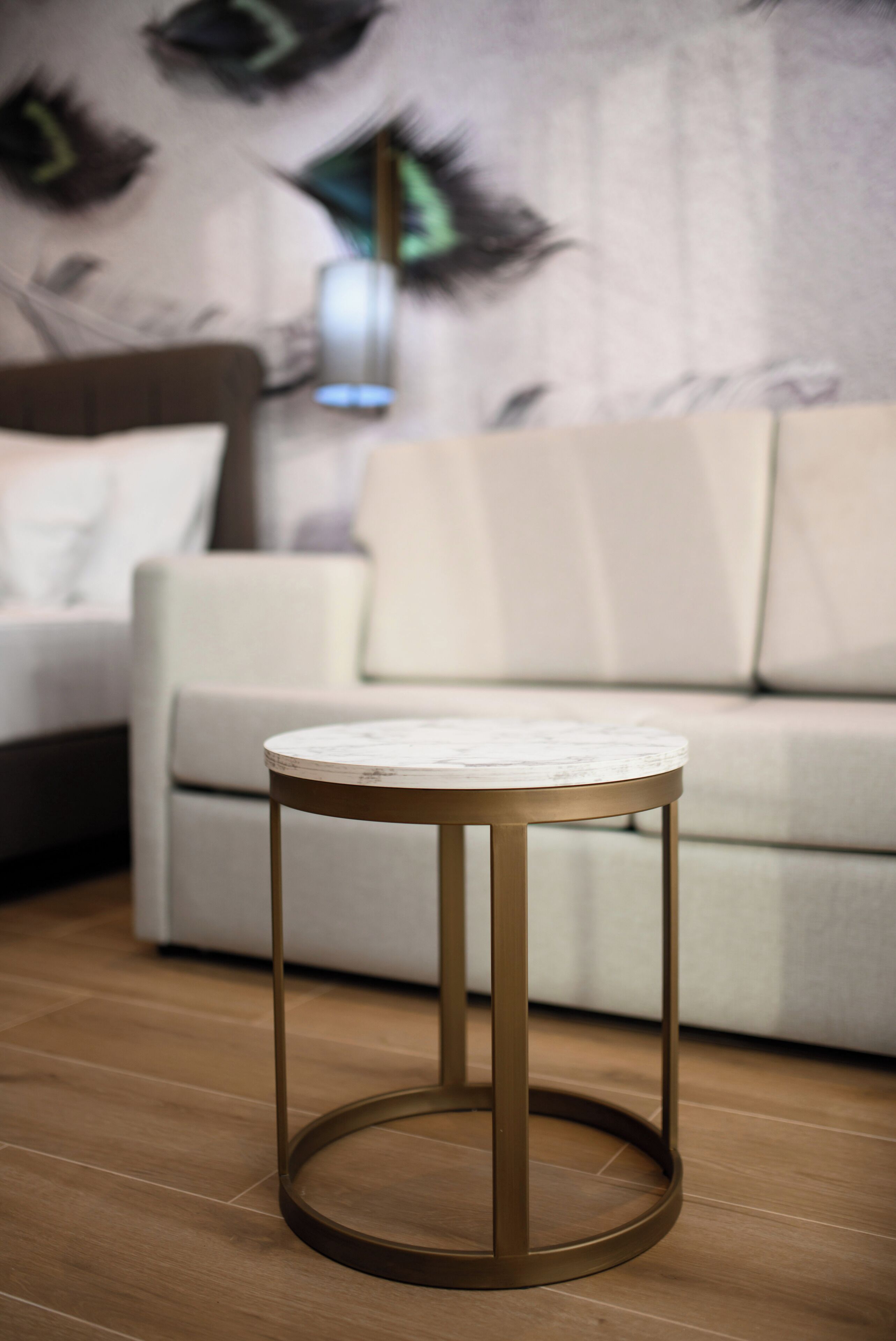 A round table with a marble top and a gold frame, placed on a wooden floor in a living room with a white sofa and a wall mural.