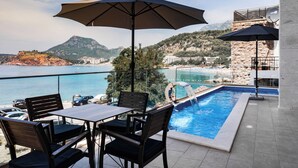 A balcony with a pool, chairs, and umbrellas overlooking a coastal area with mountains and a beach. - Hotel G Star (Sutomore)