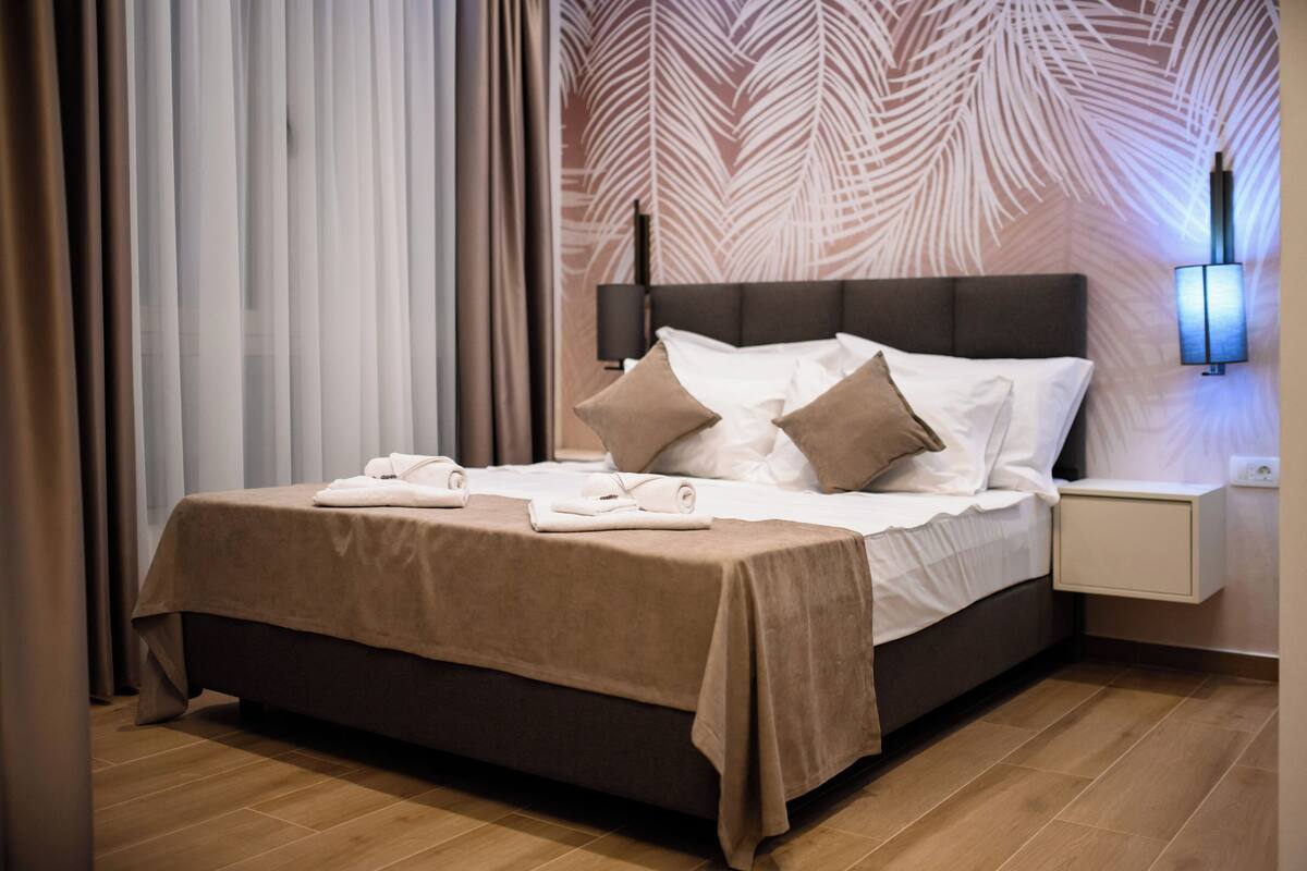 A neatly made bed with a brown bedspread and pillows, a bedside table with a lamp, and a wall with a leaf pattern.