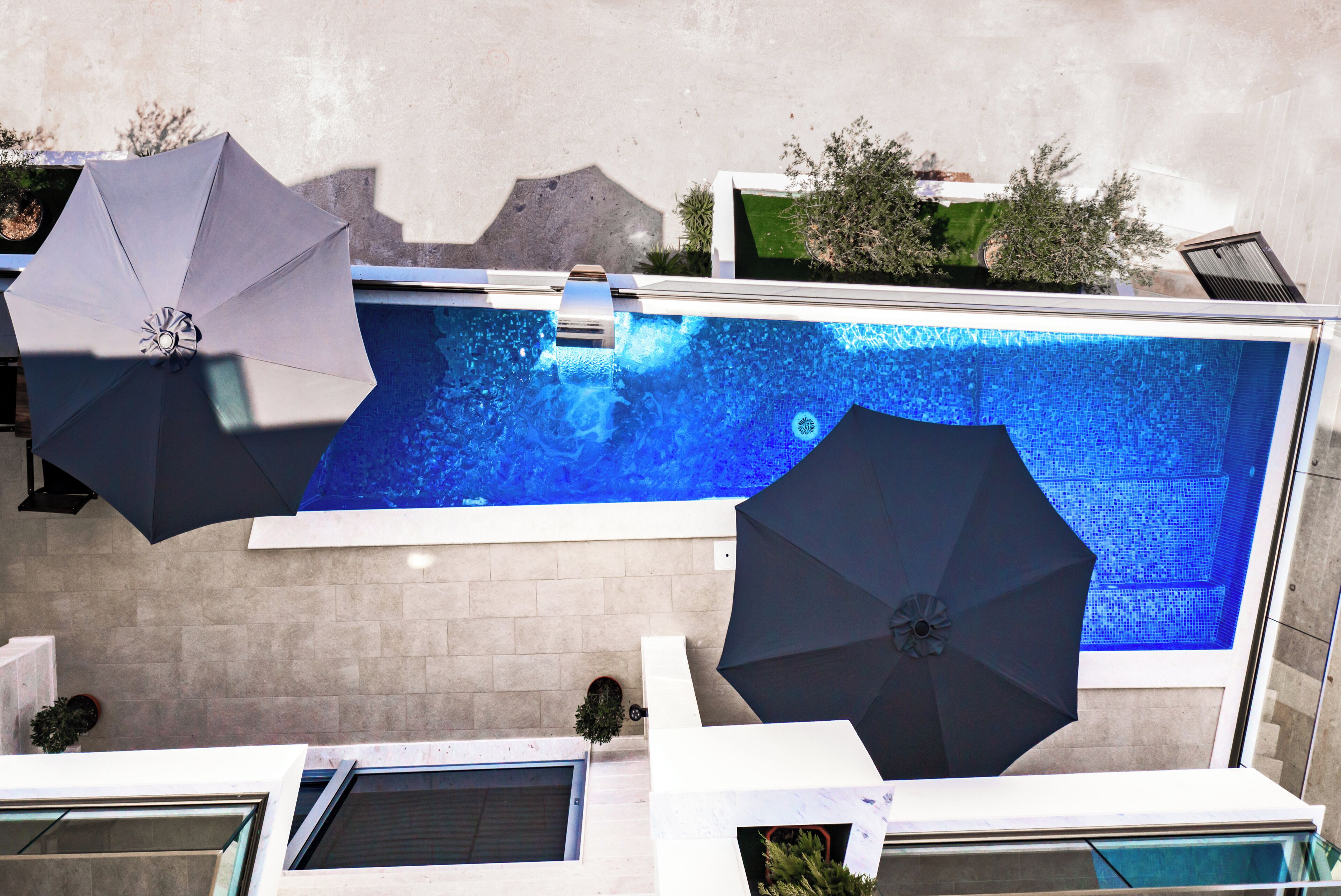 Una piscina con dos sombrillas, una pared de azulejos y una pequeña zona con plantas.